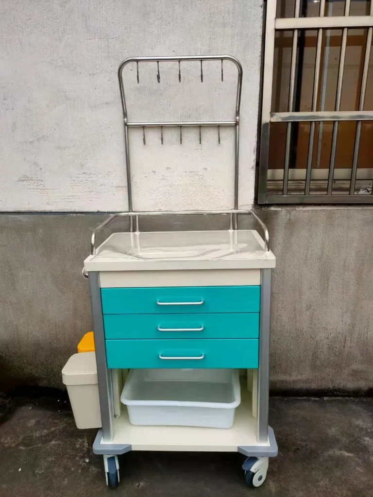 A medical equipment cart with teal drawers and a metal rack on casters placed outdoors against a wall.