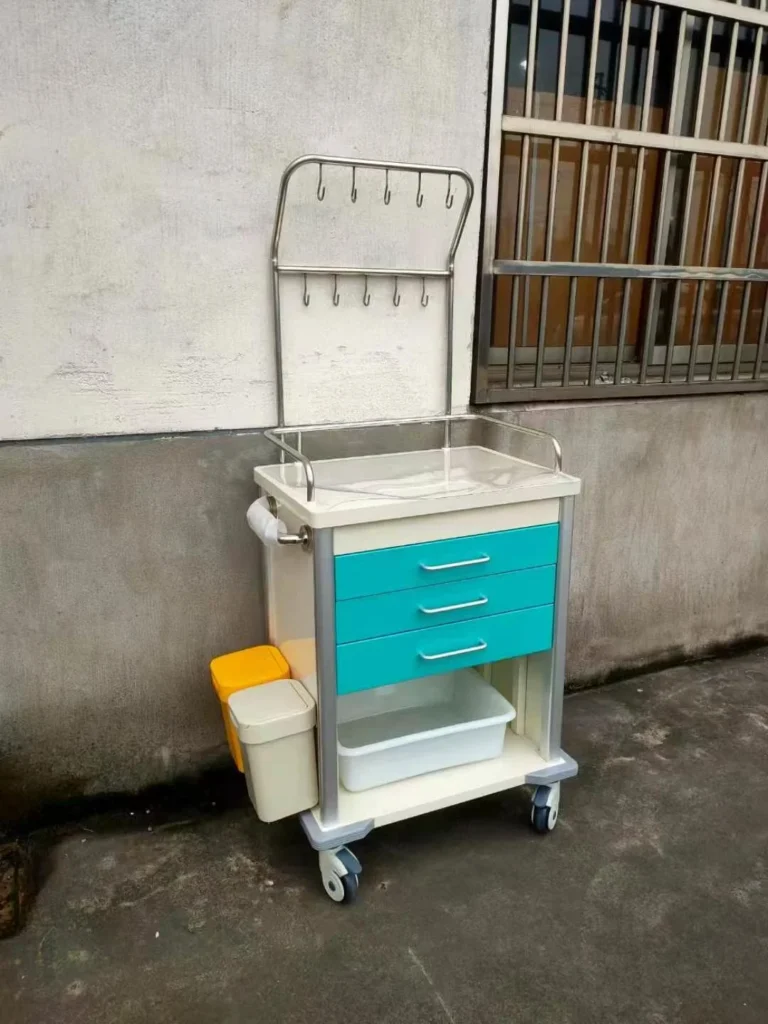 A medical storage cart with teal drawers, hooks for hanging, and attached waste bins, positioned against a wall.