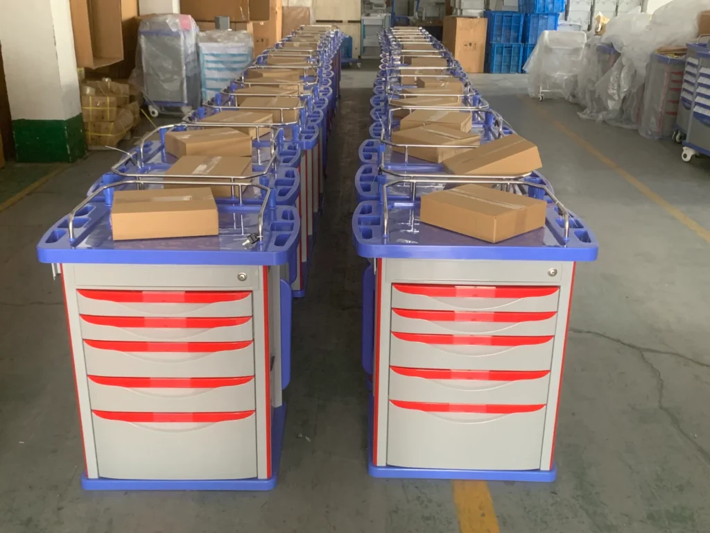 Row of medical trolleys with red drawers and blue tops, each topped with a cardboard box, organized in a warehouse setting.