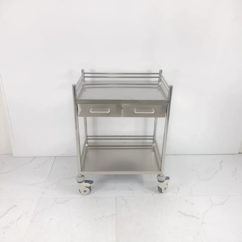 Stainless steel medical trolley with two drawers and wheels on a tiled floor against a white background.