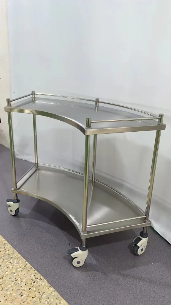 Stainless steel medical trolley with two tiers and wheels, placed indoors on a speckled floor.