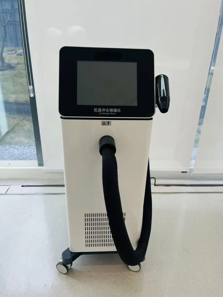 A modern cryotherapy device with a touchscreen and hose, positioned in a clinical environment near large windows.