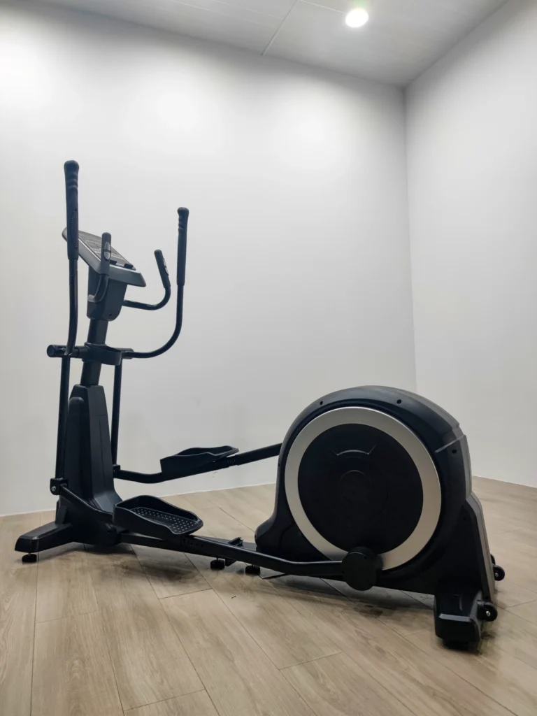 Black elliptical trainer in a minimalist gym with wood flooring and white walls, under ceiling lights.