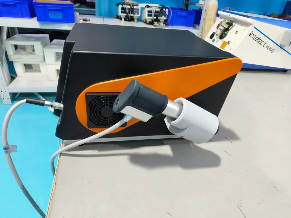 Close-up view of an industrial machine in a laboratory with blue flooring, featuring a black and orange device connected to a cable.