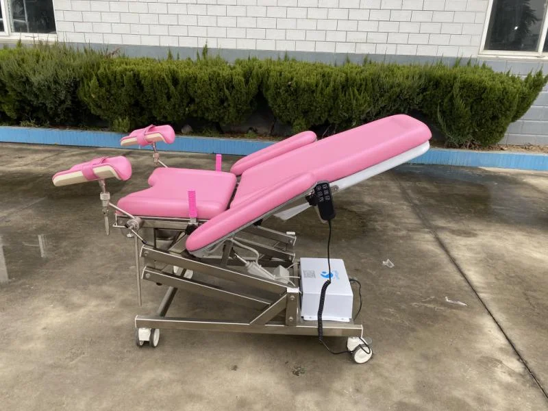 Pink medical examination chair with adjustable parts and wheels, positioned outdoors on a concrete floor with bushes in the background.