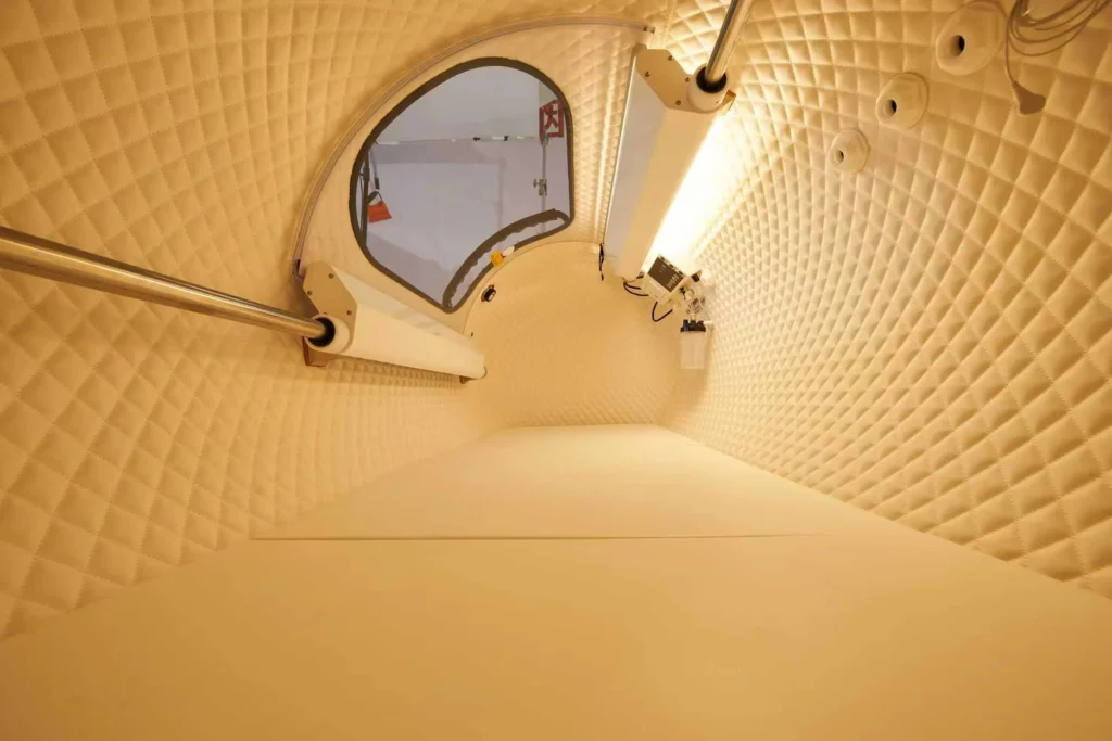 Interior of a soundproof pod featuring quilted walls, a circular window, and lighting fixtures.