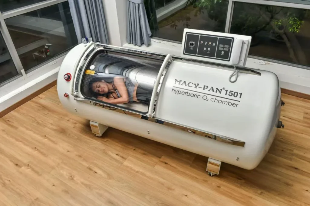 A woman resting inside a Macy-Pan 1501 hyperbaric oxygen chamber in a room with wooden flooring and large windows.