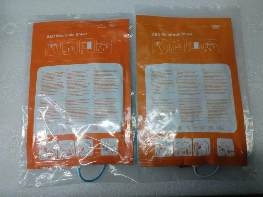 Two orange AED electrode sheet packaging bags with multilingual instructions and visual guidelines for application.