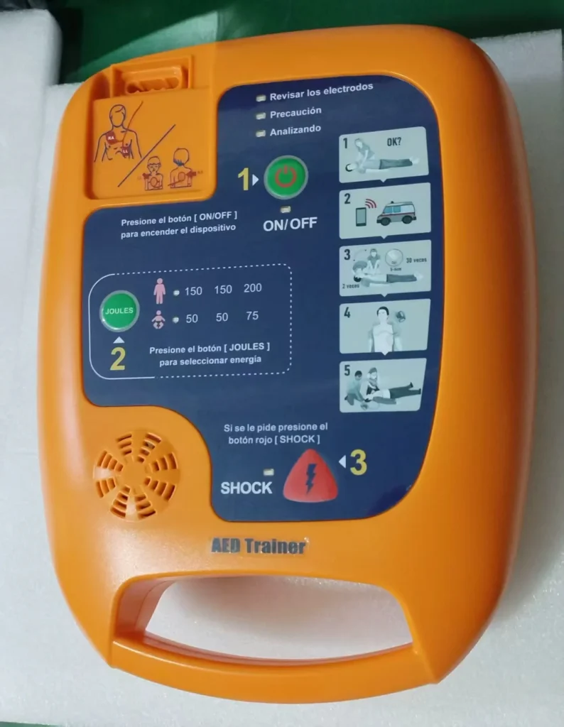 Orange AED trainer device with instructional labels on the front, featuring a power button, shock button, and visual step-by-step guidance for usage.