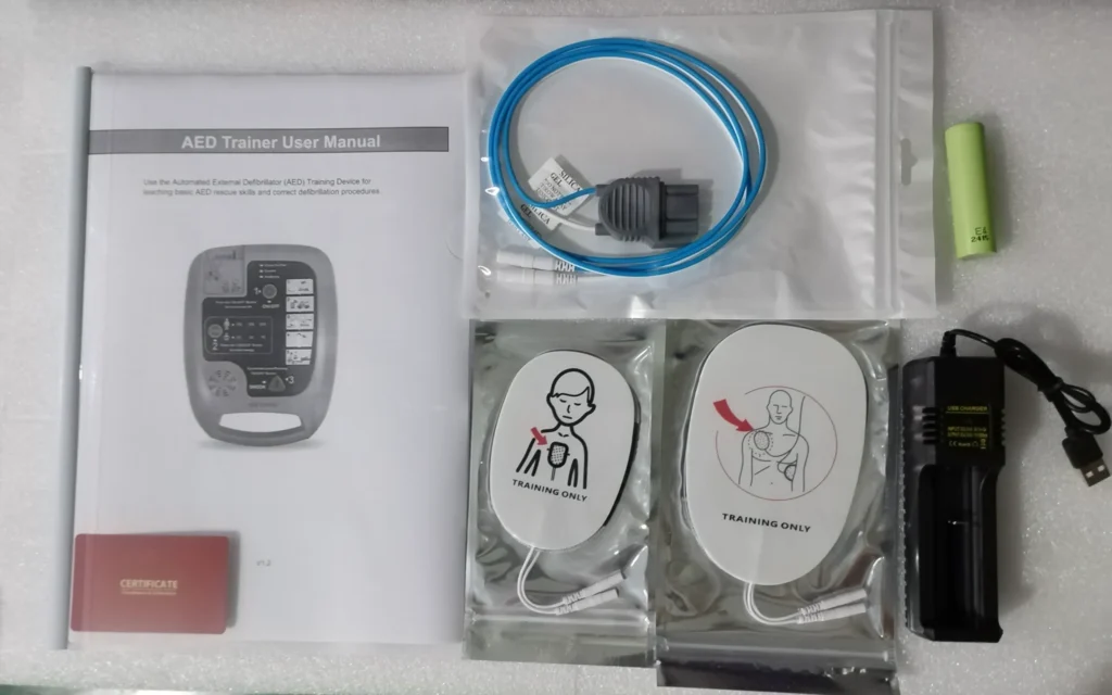 Flat lay view of an AED training kit, including a user manual, electrode pads labeled 'training only,' USB charger, battery, cables, and a certification card.