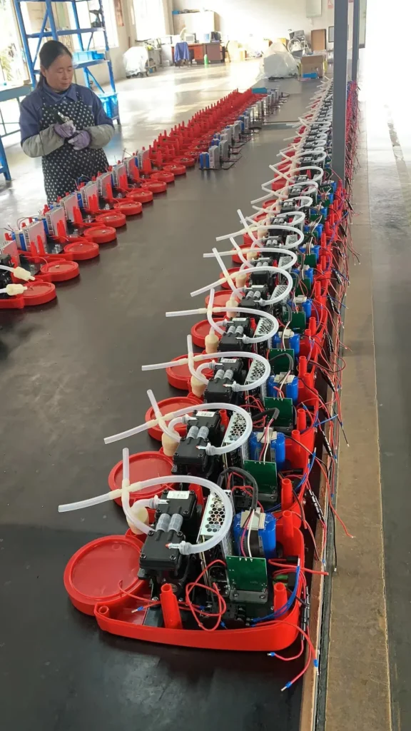 An assembly line of red electronic components with tubes and wiring, in a factory setting. A worker in gloves observes the production process.