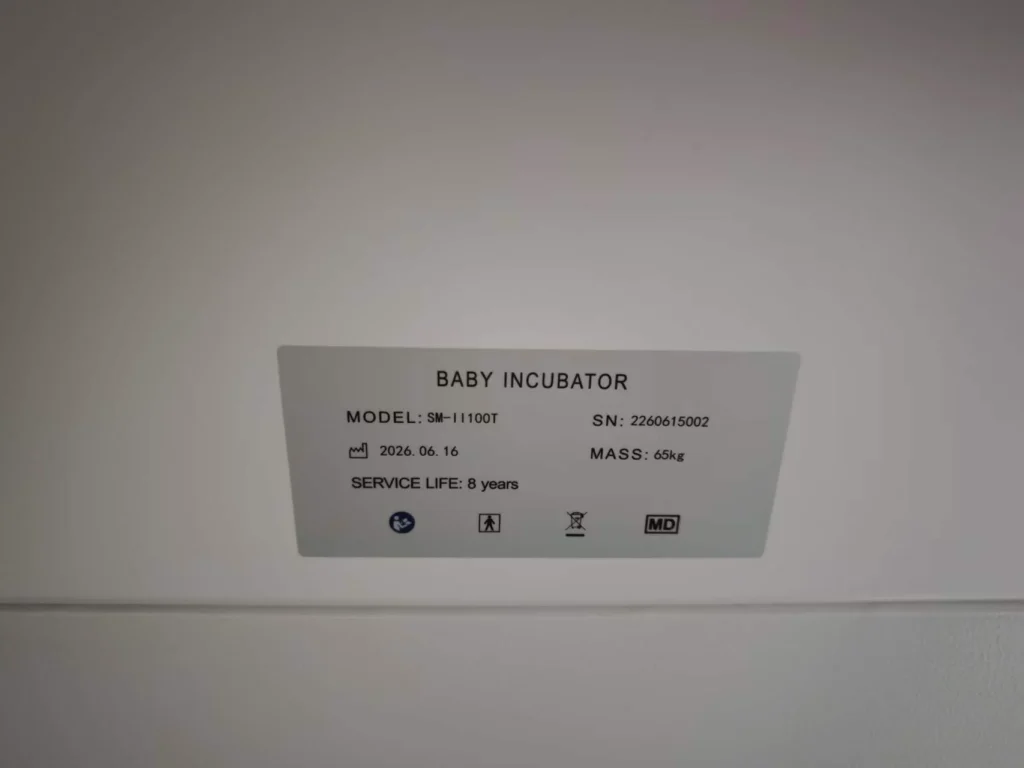 Specifications label of a baby incubator displaying model number SM-1100T, serial number 2260615002, service life of 8 years, and mass of 65 kg. Includes symbols and production date of 2026-06-16.