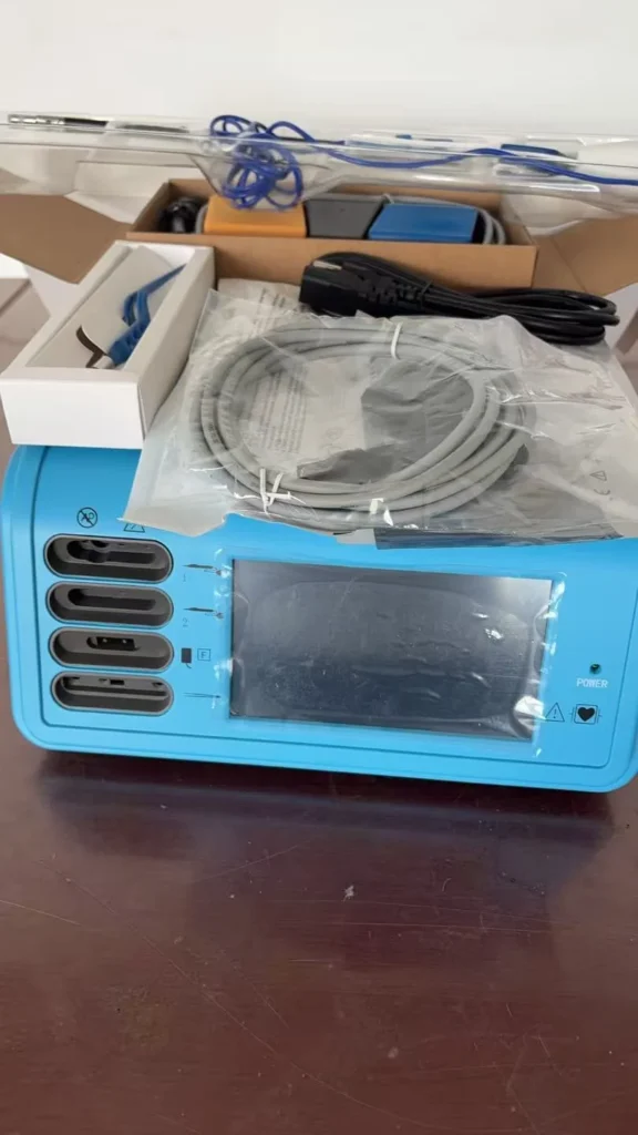 Blue medical device with a screen, ports, and packaged cables, placed on a wooden table alongside various accessories.