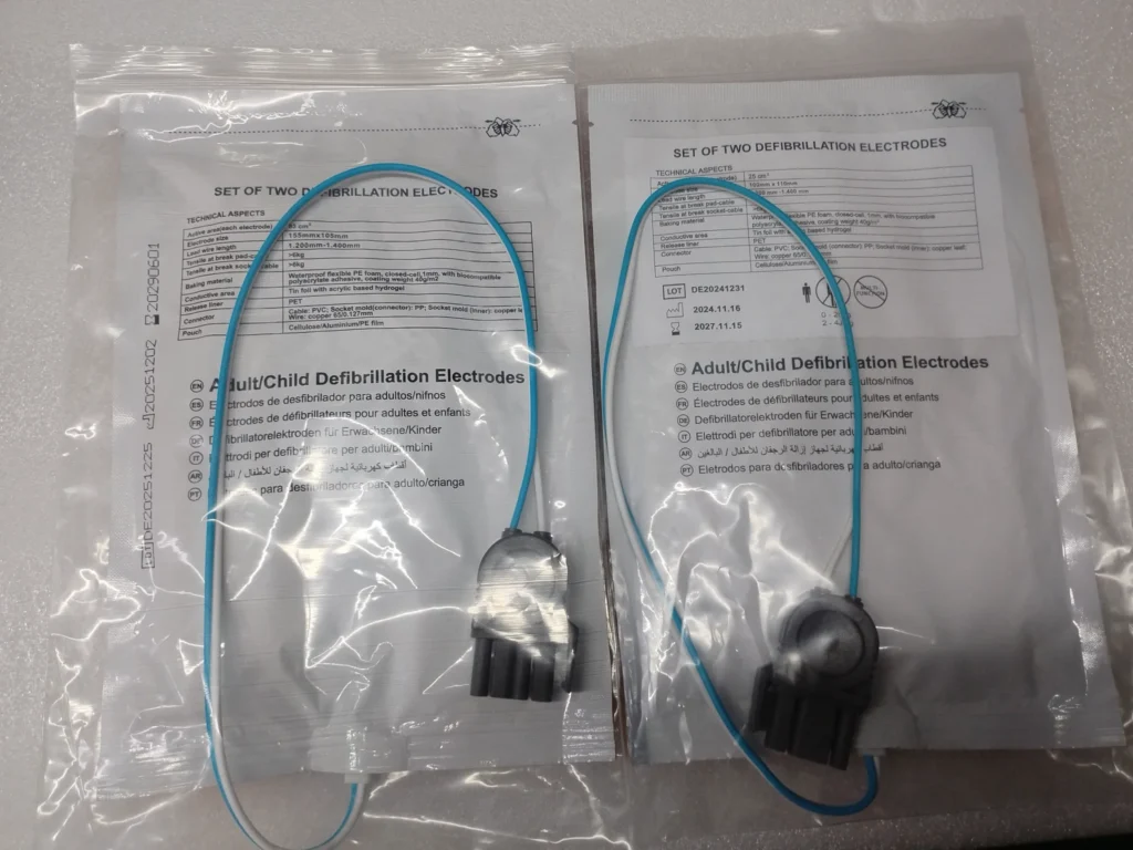 Two sets of adult and child defibrillation electrode pads in sealed transparent packaging, showing product information and blue wires.