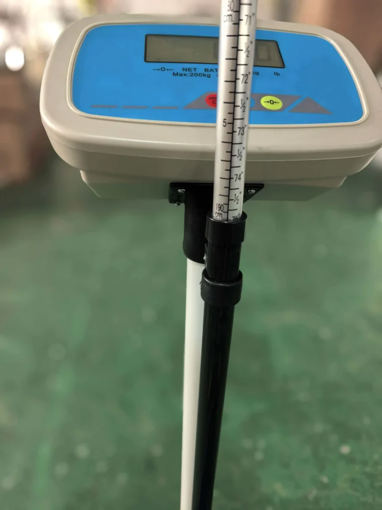 Digital weighing scale with a blue display screen and an attached height measuring rod with measurement marks, standing on a green surface.
