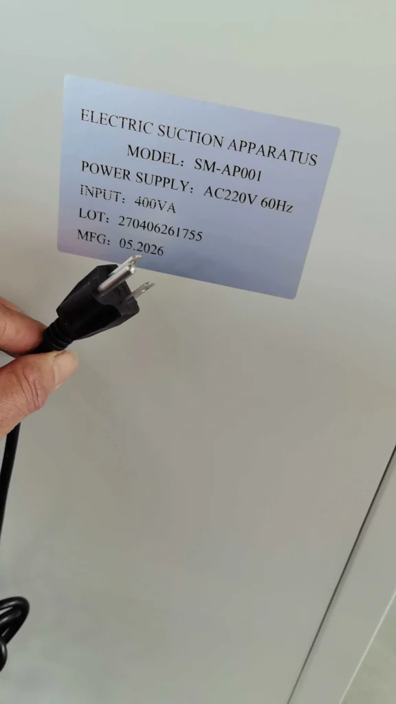 Label of an electric suction apparatus showing specifications including model SM-AP001, power supply AC220V 60Hz, input 400VA, lot number, and manufacture date, with a hand holding a black power plug.