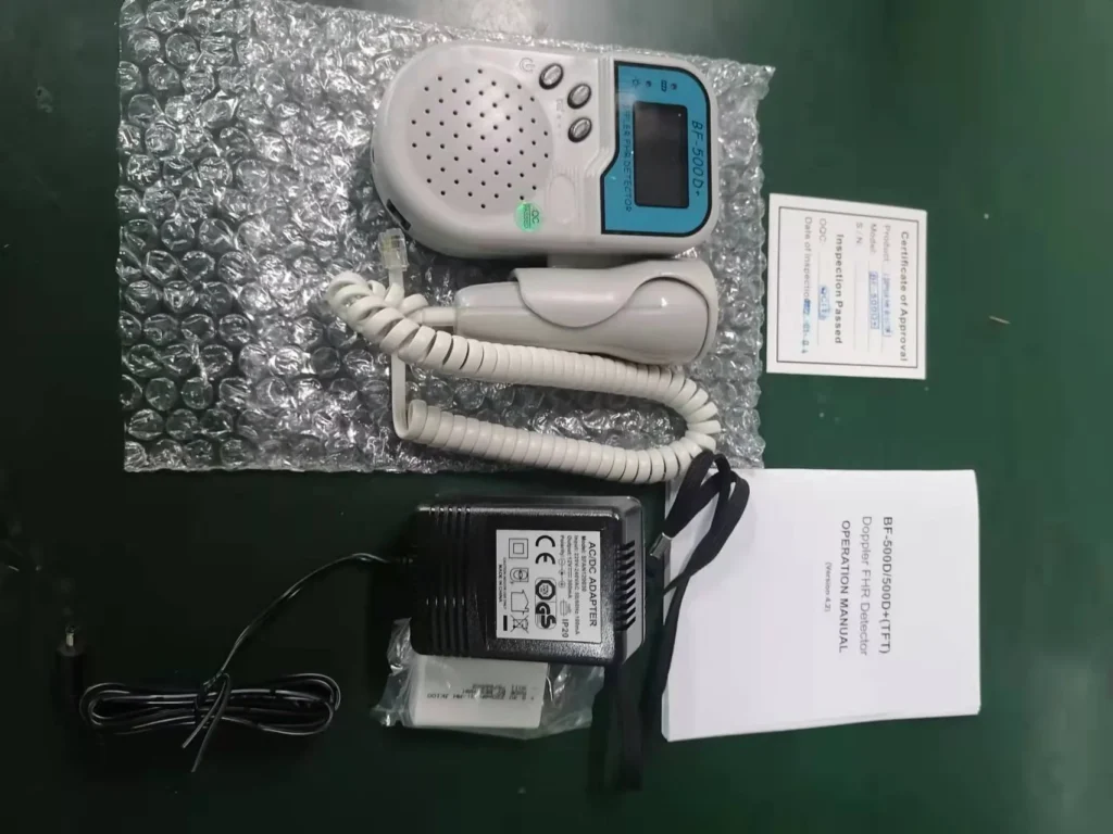 Fetal Doppler device with an LCD screen, coiled cable, power adapter, user manual, and inspection certificate on a bubble wrap surface.