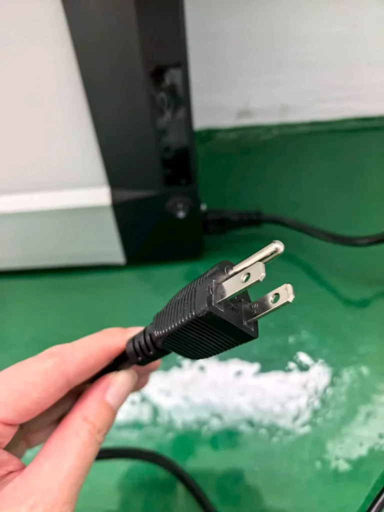Close-up of a hand holding a black power cord with a two-prong plug, with a green surface and a piece of electronic equipment in the background.