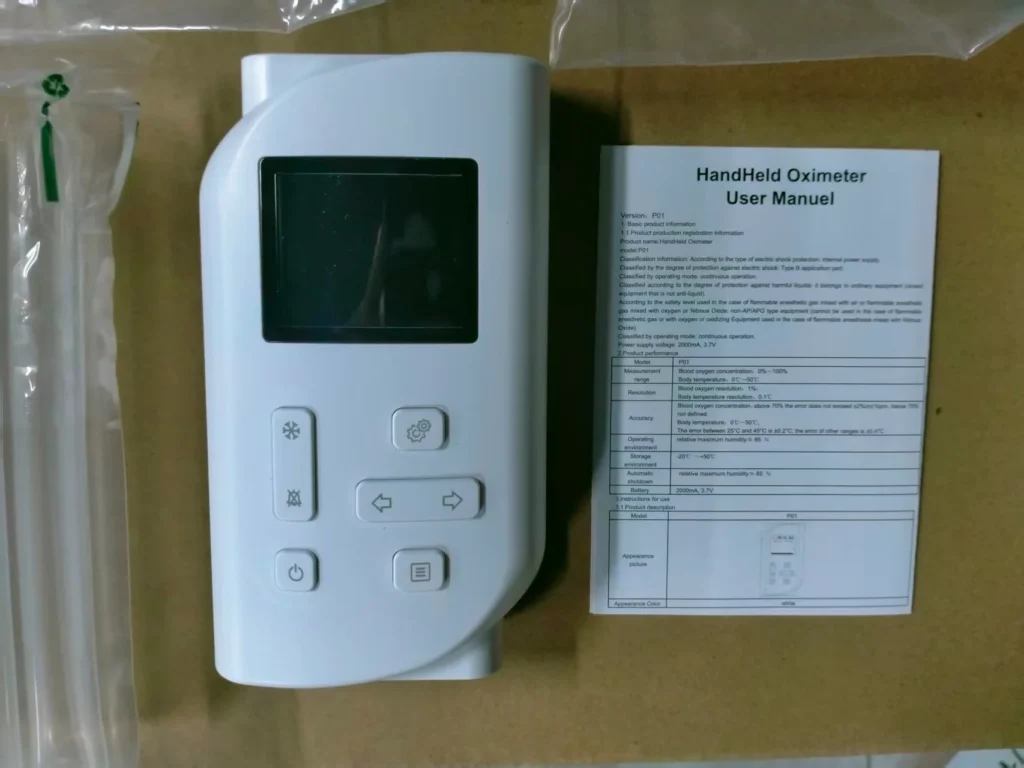 A handheld oximeter device placed alongside its user manual on a light brown surface, with clear plastic tubing nearby.