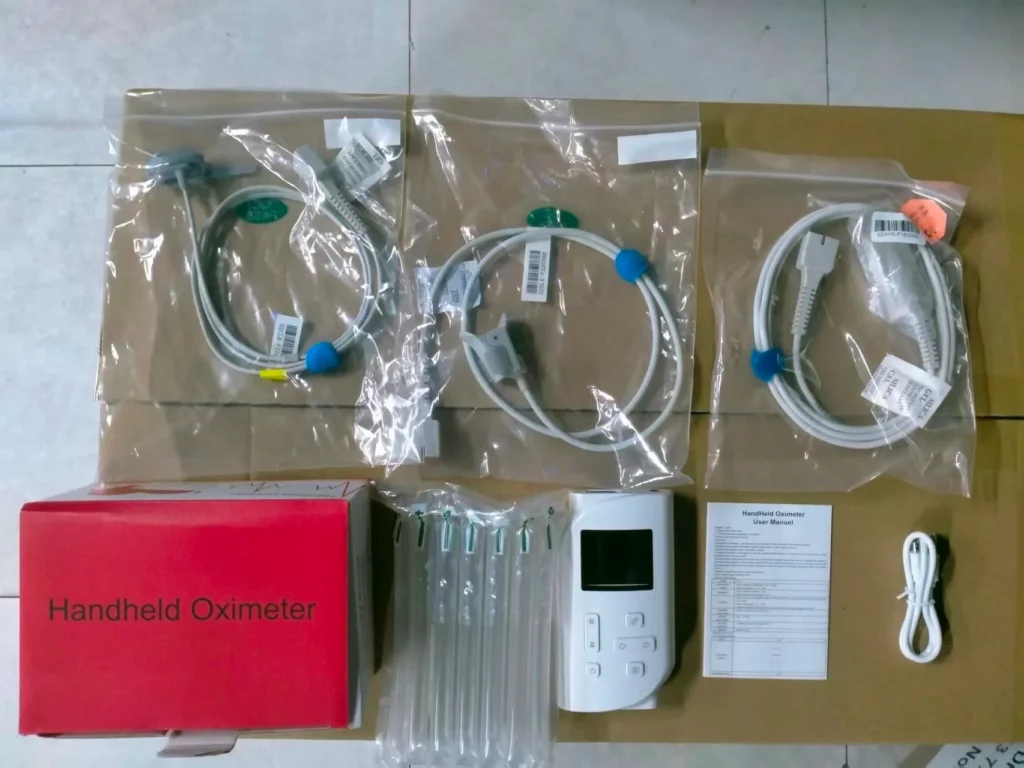 Handheld oximeter device with cables, user manual, and other accessories arranged on a flat surface in their packaging.