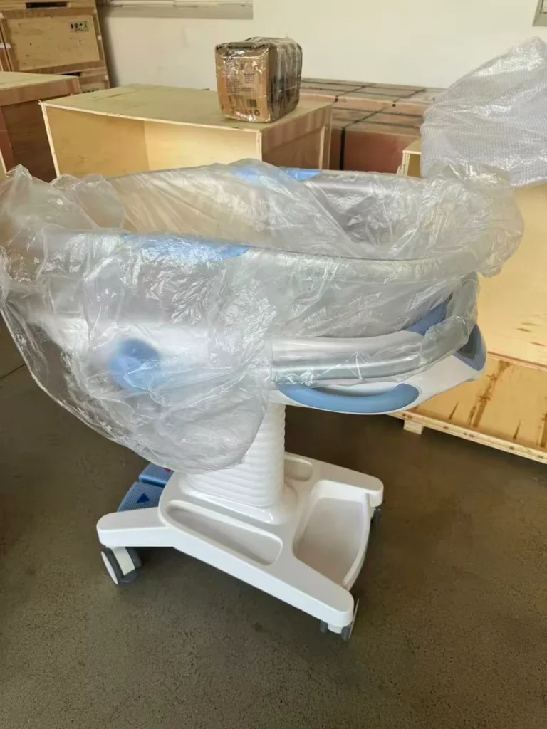 A hospital baby bassinet covered with protective plastic, placed in a warehouse, surrounded by wooden crates and boxes.