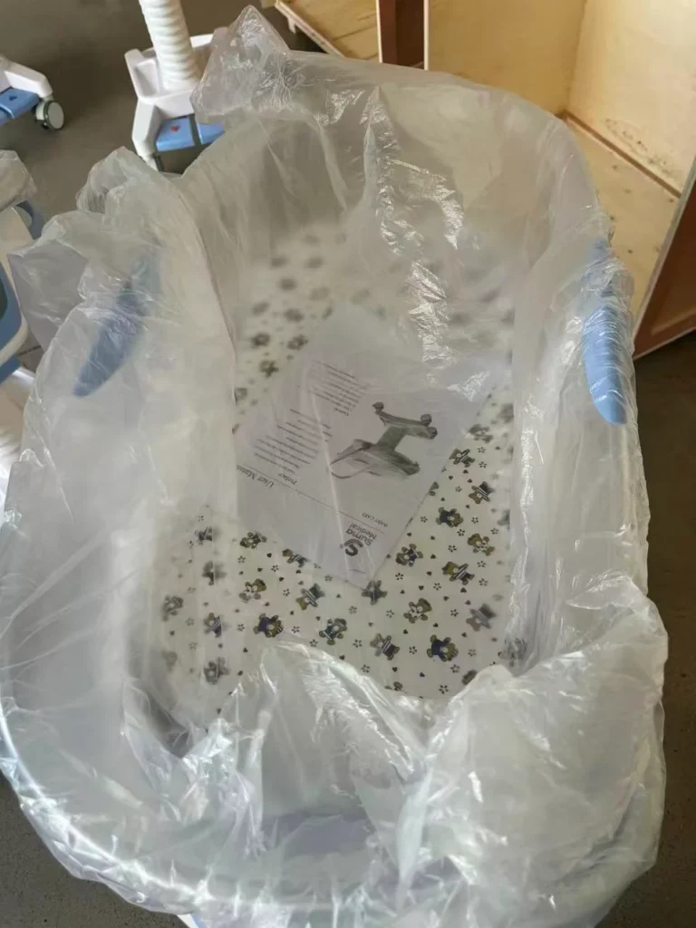A hospital baby bassinet with a plastic cover, floral bedding, and an instruction sheet placed on the mattress, located indoors.