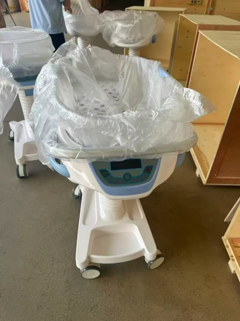 Hospital infant bassinets on wheels covered in protective plastic, positioned in a warehouse or preparation area.
