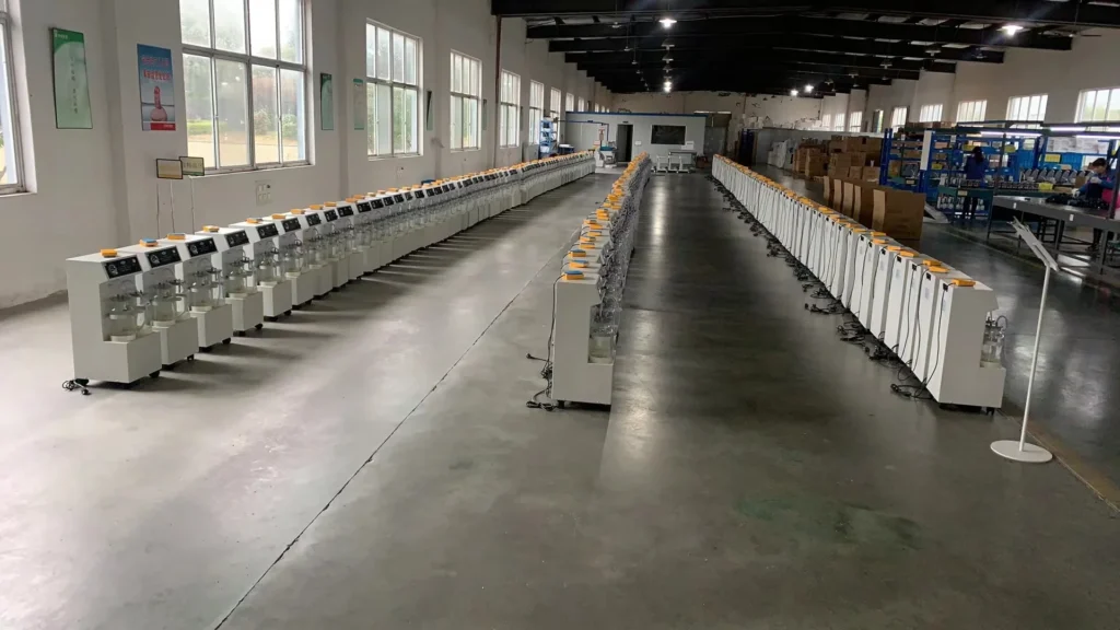 Long rows of identical machines arranged in an industrial assembly line inside a well-lit factory with large windows and open space.
