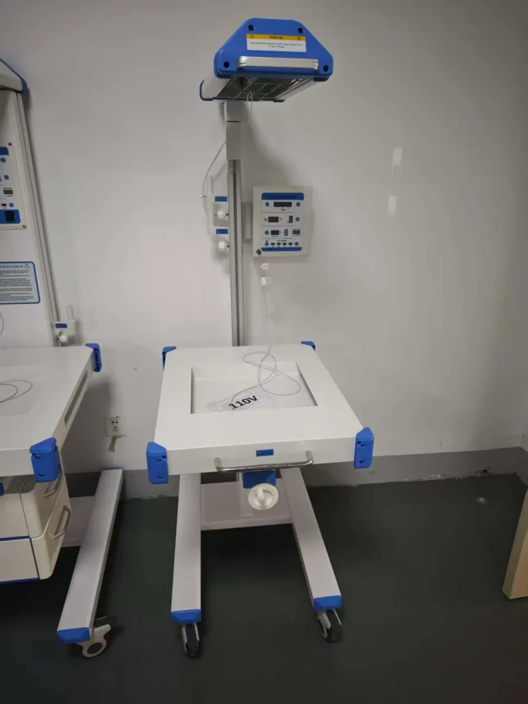 A hospital-grade infant radiant warmer with blue and white accents, mounted on wheels, equipped with a control panel and overhead heating unit.