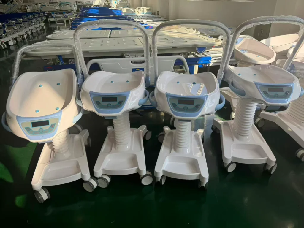 Four white infant warmers with digital controls on wheels lined up in a medical facility with hospital beds in the background.