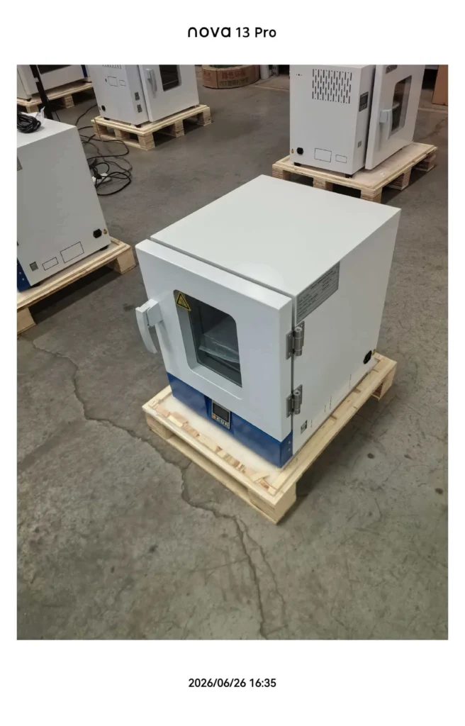 A compact laboratory oven placed on a wooden pallet in an industrial setting. Other equipment is visible in the background.