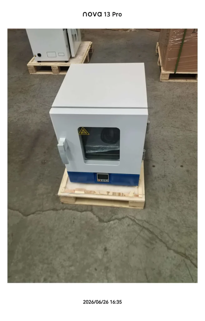 A laboratory oven with a glass window and control panel, placed on a wooden pallet in an industrial setting.