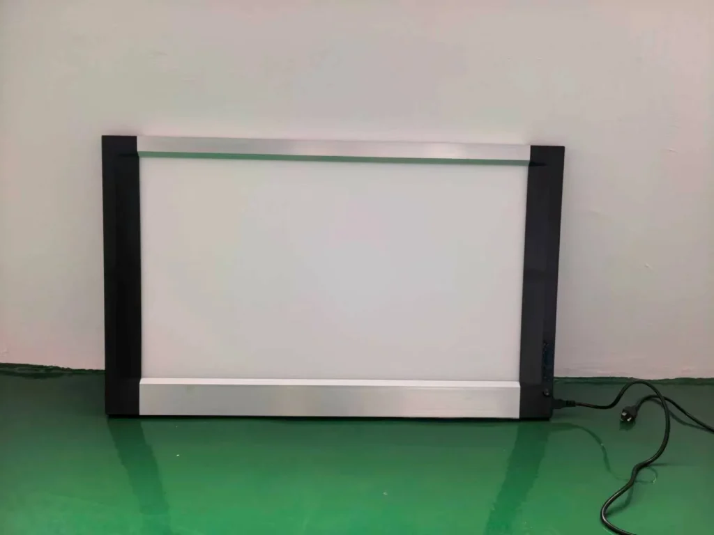 Rectangular LED light panel with aluminum frame and black side panels resting on a green floor, with a visible power cord.