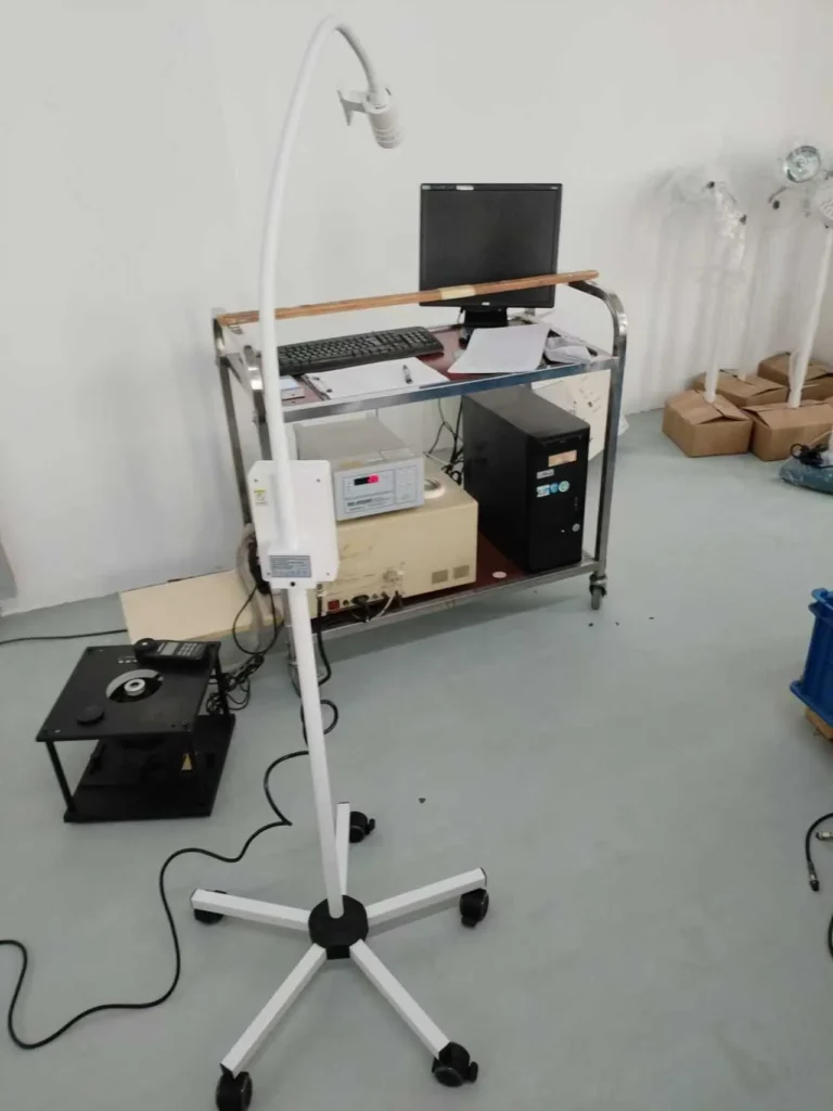 Medical examination lamp on a wheeled stand next to a workstation with a computer, medical devices, and cables in a clinical setting.