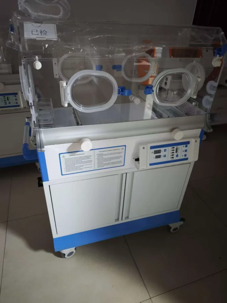 A medical infant incubator with transparent walls and control panel on wheels, placed indoors with a tiled floor.