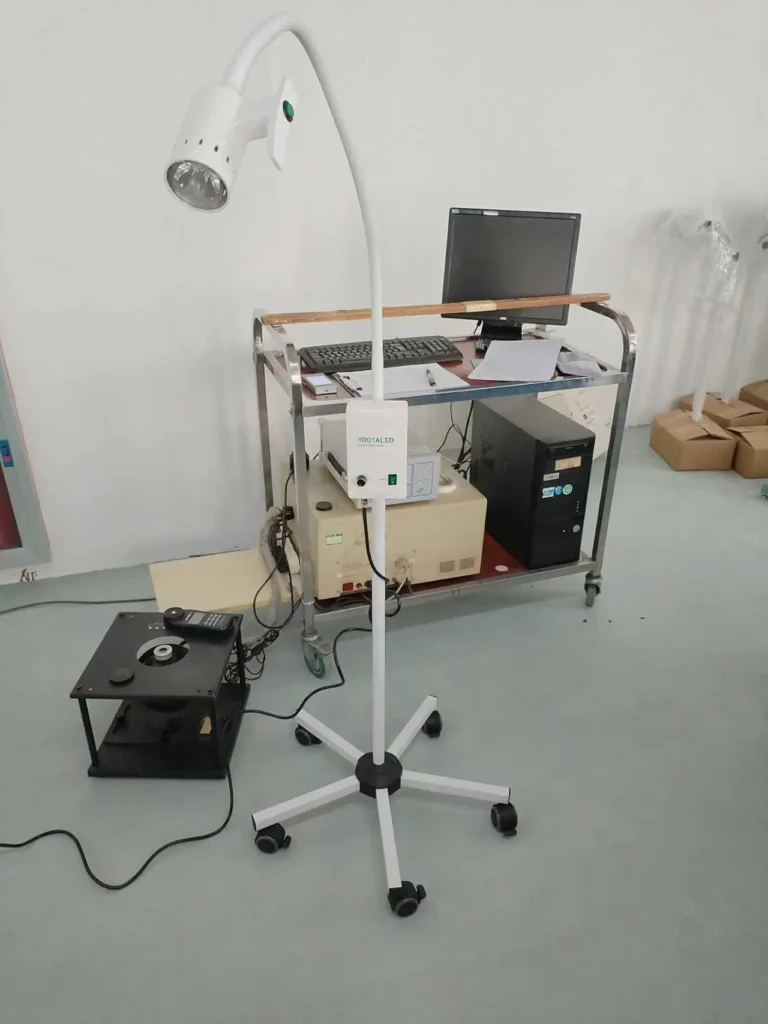 Mobile LED lamp with adjustable arm and wheeled base placed next to medical equipment and a computer on a wheeled cart in a clinical setting.