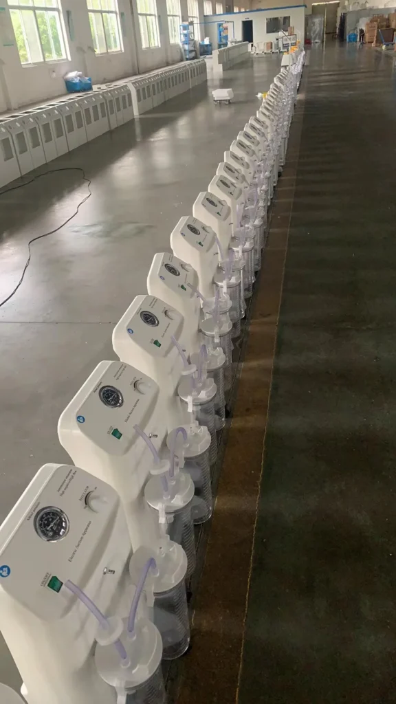 Line of white electrical medical suction machines with clear collection jars positioned in a factory setting with large windows.