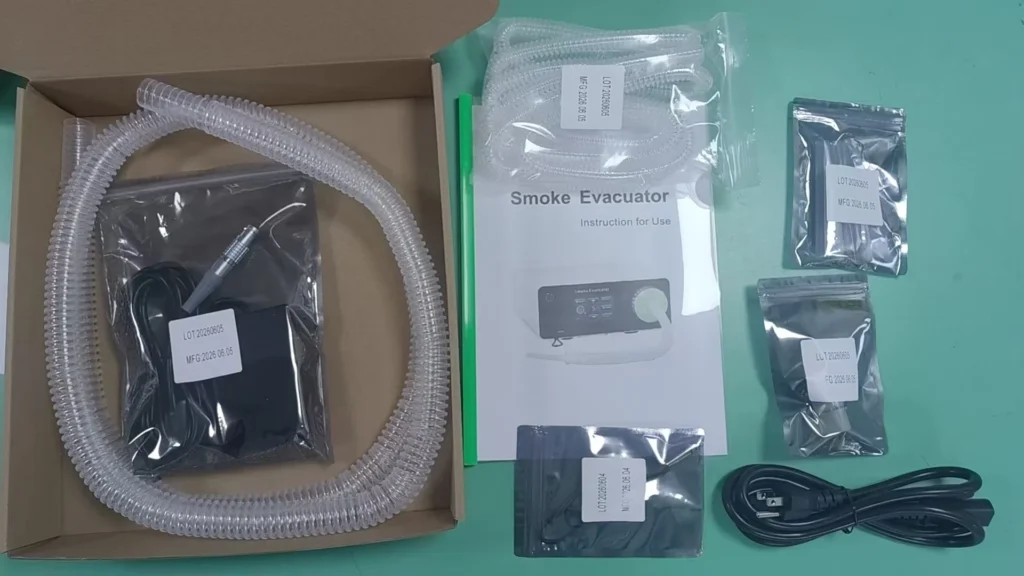 Unboxed components of a smoke evacuator kit including plastic hose, power cable, instructional manual, and various sealed parts on a green surface.
