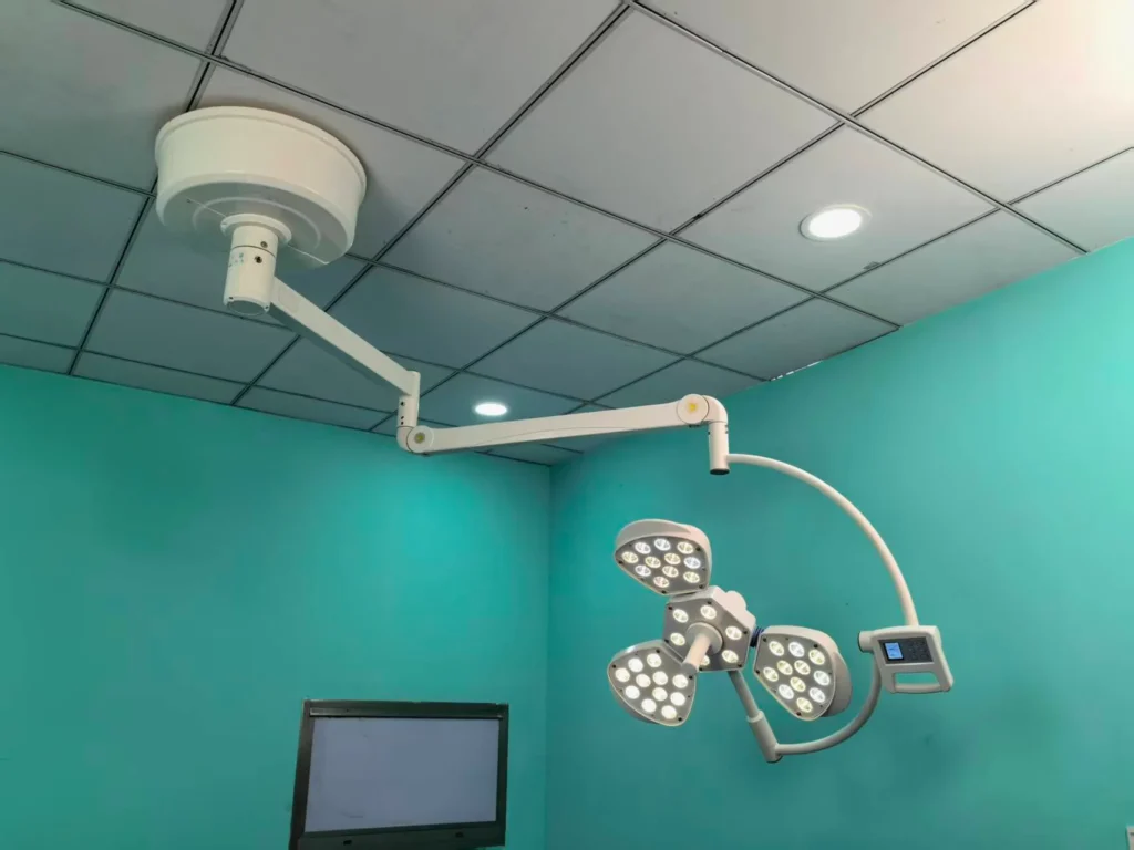 Ceiling-mounted surgical LED light with adjustable arms installed in a modern operating room with teal walls and tiled ceiling.