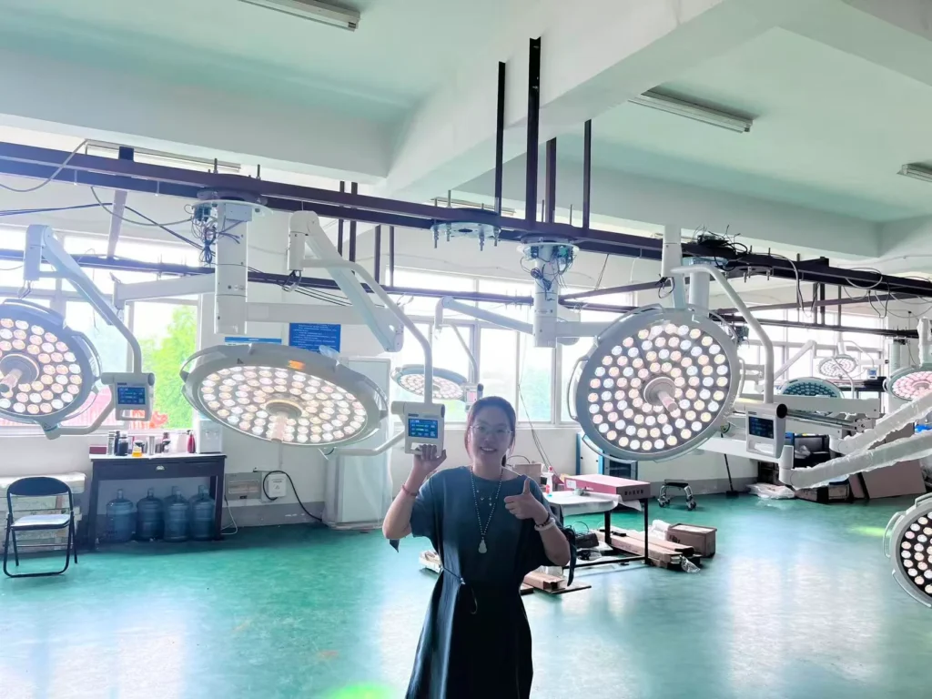 A person in a factory setting standing beneath large LED surgical lights, giving a thumbs-up. Several surgical lights are hanging from the ceiling.
