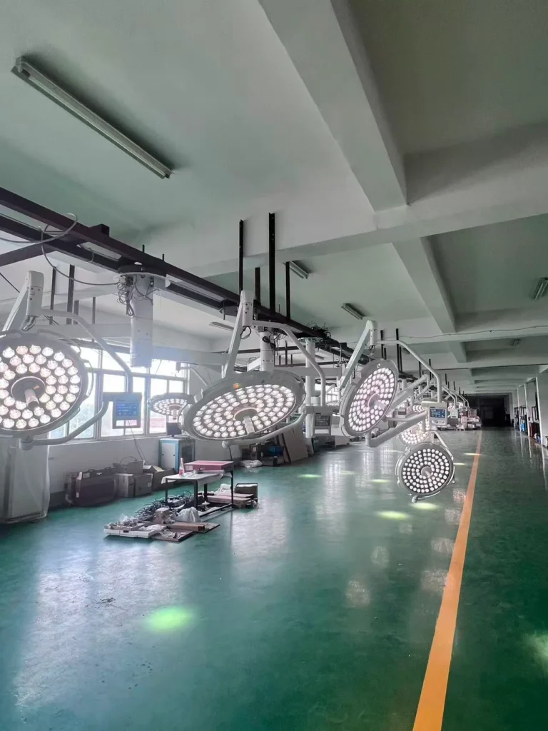 A row of surgical lights hanging on racks in a spacious production facility with large windows and green flooring.