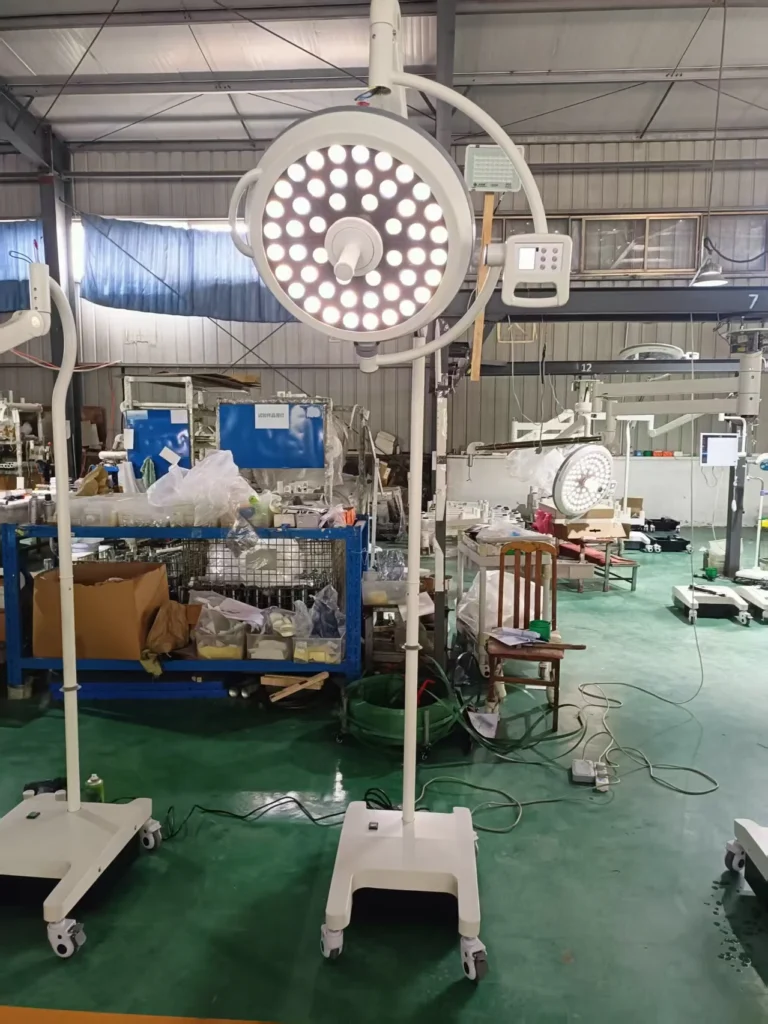 A large surgical operating light on a wheeled stand in a manufacturing workshop, surrounded by industrial equipment.