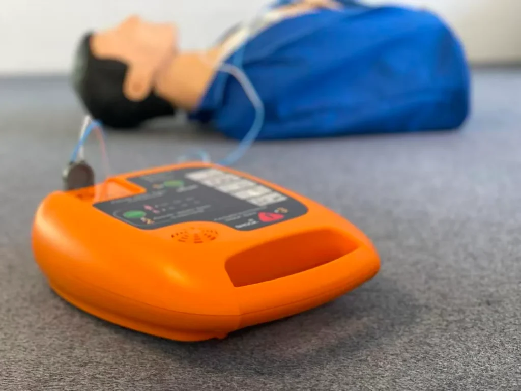 An orange automatic external defibrillator (AED) on a carpeted floor next to a practice mannequin used for CPR training.