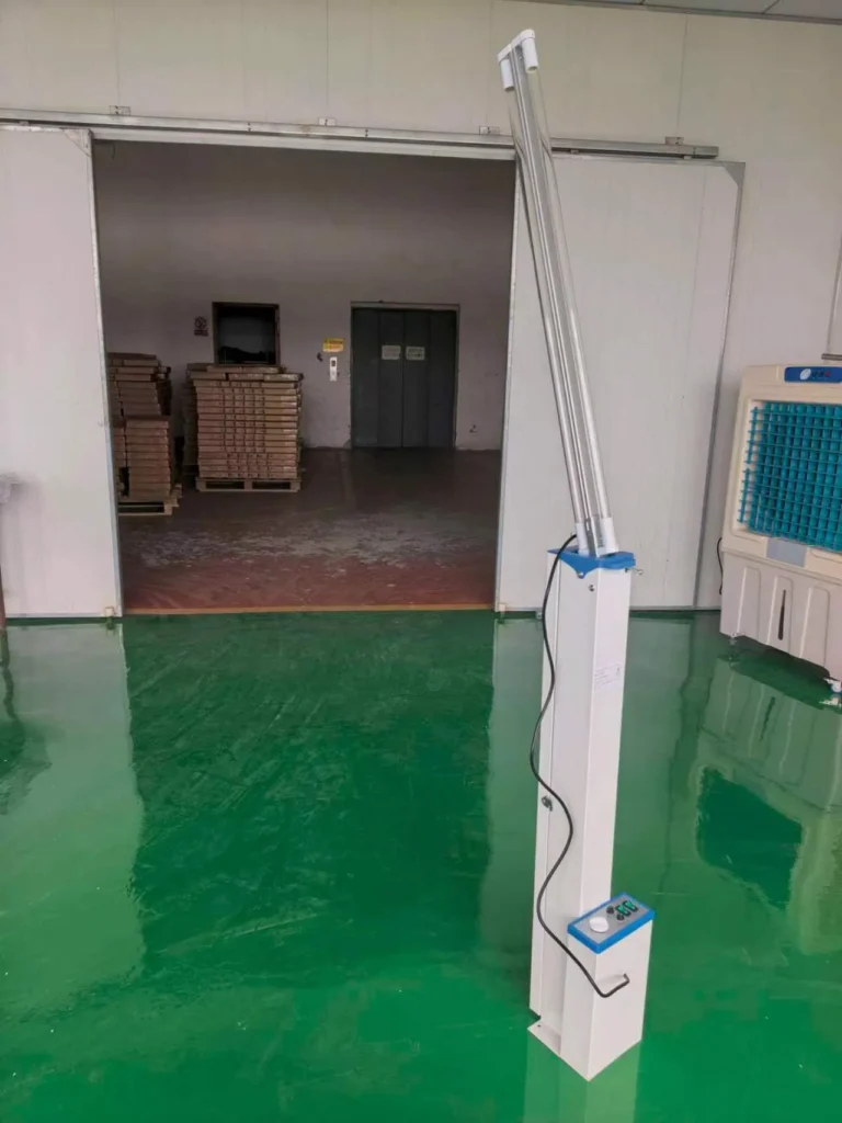 A UV sterilization lamp with a tall white base and control panel standing on a shiny green floor in a factory setting, with open doors revealing stacked pallets and an industrial area.