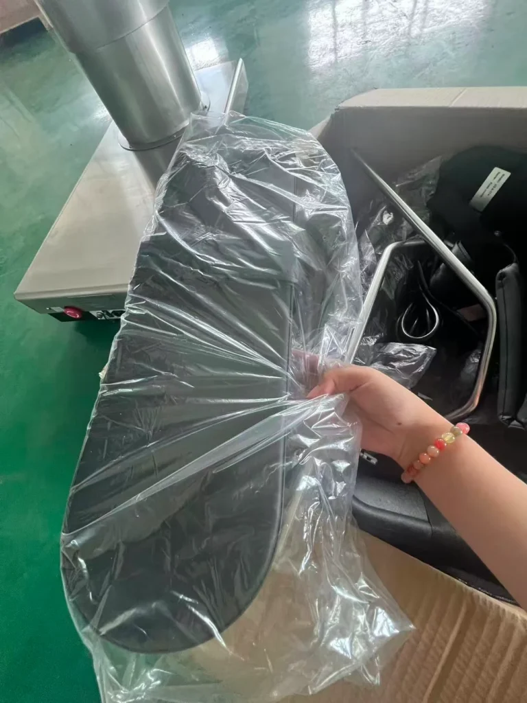 A hand holding a plastic-wrapped black equipment piece, with a stainless steel machine and a cardboard box visible in the background.