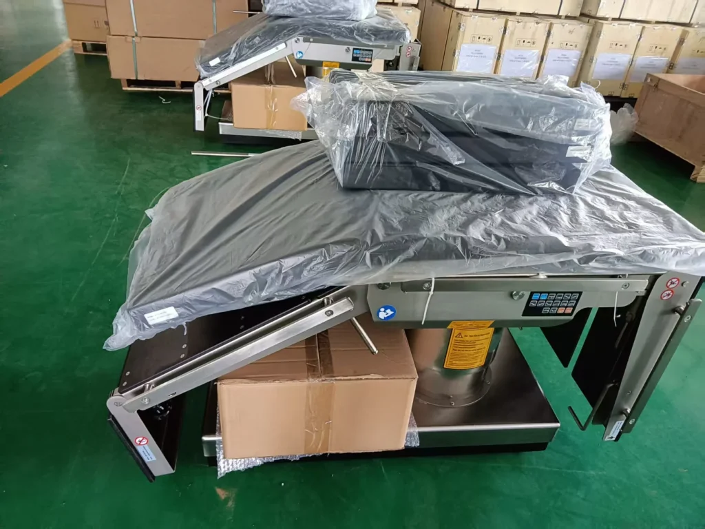 Industrial weighing machine wrapped in plastic and partially packaged, placed in a warehouse with green flooring and stacked boxes.