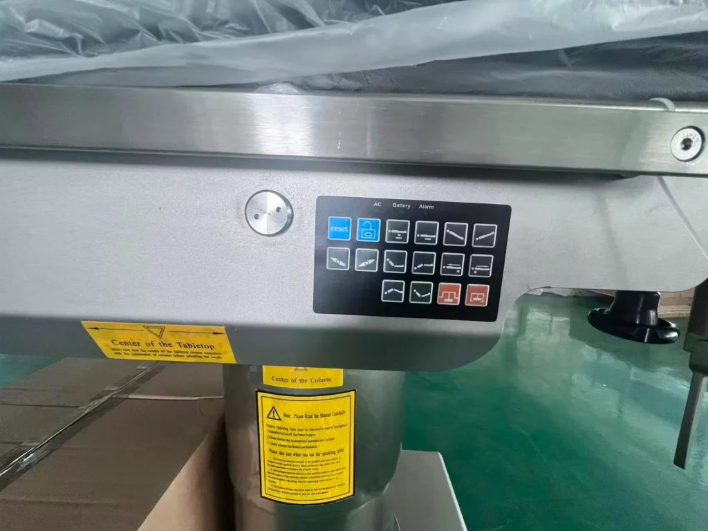 Close-up view of a medical operating table control panel with various buttons and warning labels on a metallic surface.