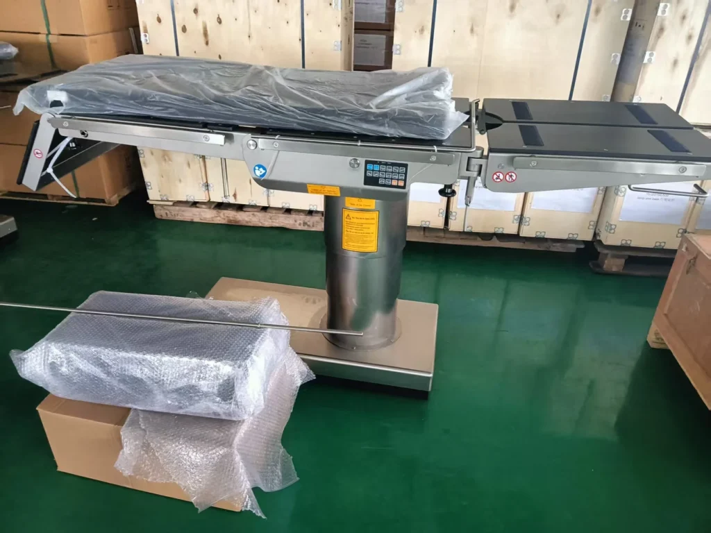 A surgical operating table on a green floor, partially assembled and covered with protective wrap, with boxed accessories nearby.
